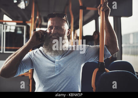 Mature man with a long beard laughing while standing on a bus talking on his phone during his morning commute Stock Photo