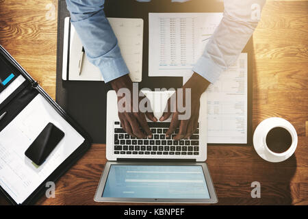 High angle of an African businessman sitting at his desk in an office busy working online with a laptop and going over documents Stock Photo