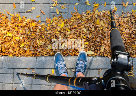 View from a tripod to the feet of a photographer taking pictures of the texture of paving stones in the autumn season. Blue Sneakers on a Gray Paving  Stock Photo