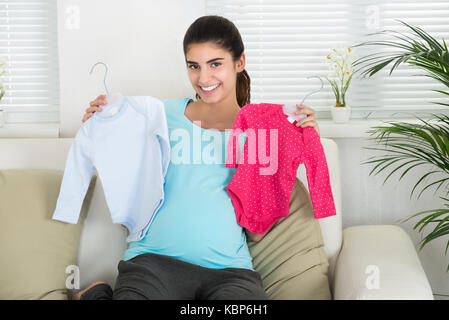 Portrait of happy pregnant woman holding baby clothes while sitting on sofa at home Stock Photo