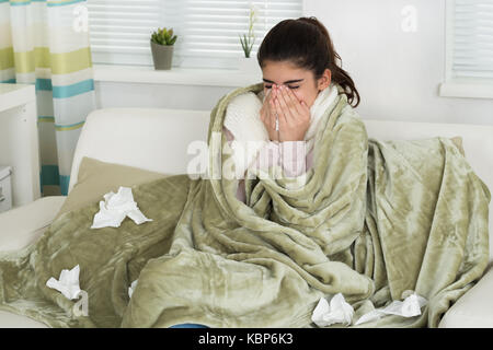Sick young woman covered with blanket blowing nose while sitting on sofa at home Stock Photo