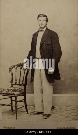 American archive monochrome studio portrait photograph of young man wearing light coloured shirt and trousers and long dark jacket standing next to upholstered dining room chair, named as Isac Gage, taken in late 19th century by Webster Photographers, Binghamton, NY, USA Stock Photo