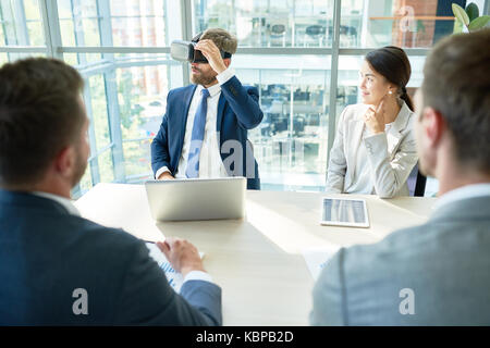 Bearded middle-aged entrepreneur using VR headset while having working meeting with colleagues concerning promising start-up project, interior of mode Stock Photo