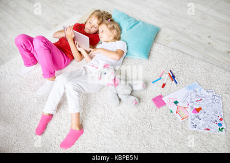 High angle portrait of two sisters using digital tablet lying on floor on thick plush carpet in cozy living room while alone at home Stock Photo