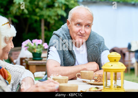 Waist-up portrait of handsome senior man with deep blue eyes looking away pensively while sitting at table with friends and celebrating momentous even Stock Photo