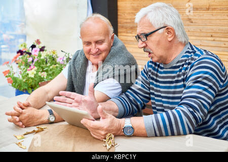 Two handsome senior friends using digital tablet while sitting at outdoor cafe table and waiting for their order Stock Photo