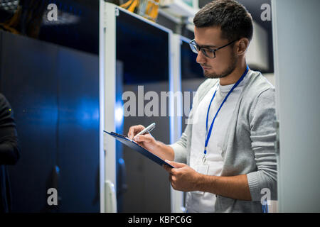 Side view portrait of young network engineer writing data on clipboard while working with servers in data center Stock Photo