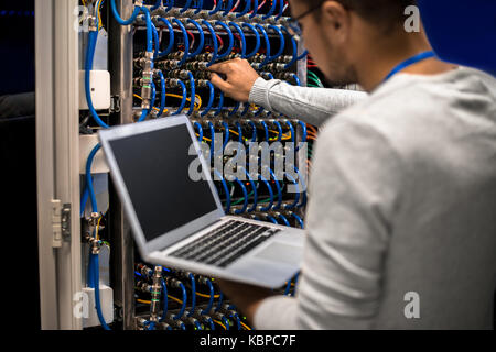 Back view portrait of young man working with supercomputer connecting blade server cables and  checking data on laptop Stock Photo