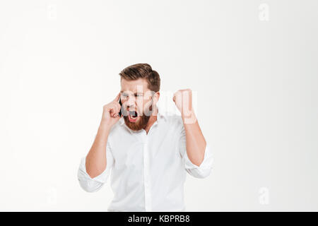 Image of screaming angry young bearded emotional man standing over white wall background isolated. Looking aside while talking by phone. Stock Photo