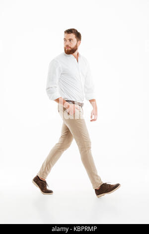 Picture of handsome young bearded man walking over white wall background isolated. Looking aside. Stock Photo