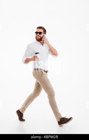 Picture of handsome young bearded man walking over white wall background isolated drinking coffee. Looking aside talking by phone. Stock Photo