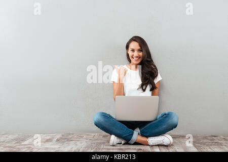 Happy smiling asian woman working on laptop computer while sitting on the floor with legs crossed and pointing finger away isolated over gray backgrou