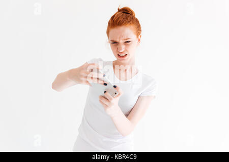 Confused ginger woman in t-shirt playing on her smartphone over white background Stock Photo