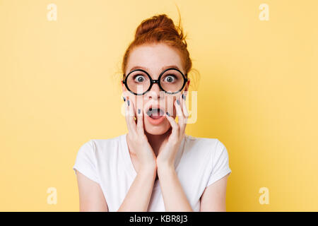 Close up picture of shocked ginger woman in eyeglasses holding her cheeks and looking at the camera Stock Photo