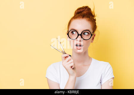 Surprised ginger woman in eyeglasses holding dividers and looking away over yellow background Stock Photo