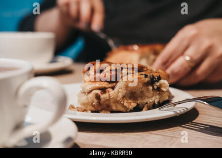 A freshly baked Chelsea buns on a plate in a cafe, cutlery and cups of tea around, female hands holds fork Stock Photo