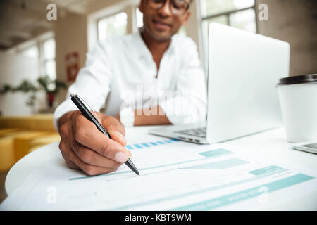 Cropped image of young african man sitting coworking with laptop computer writing notes in documents. Stock Photo