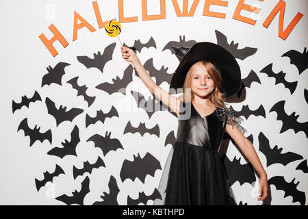 Cute little girl dressed in halloween costume holding lollipop while posing with bats on a background Stock Photo