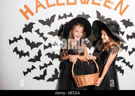 Couple of two cheerful little girls dressed in halloween costumes holding basket full with candy while posing with bats on a background Stock Photo