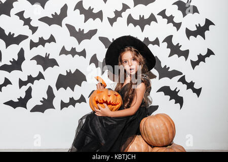 Pretty cheerful little girl dressed in halloween costume sitting with pumpkins and bats on a background Stock Photo