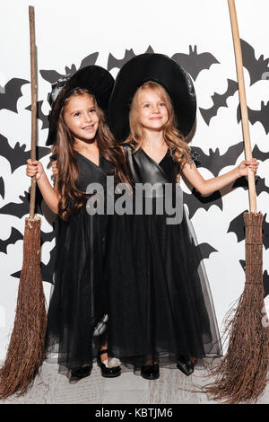 Couple of two funny little girls dressed in halloween costumes holding brooms and posing with bats on a background Stock Photo