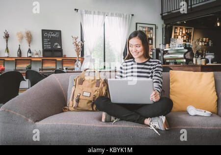 Asian teenager girl using laptop computer and listening music on sofa with happy smiling face at coffee shop,Digital age lifestyle. Stock Photo