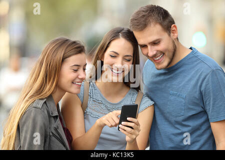 Happy group of three friends using a smart phone standing outdoors on the street Stock Photo