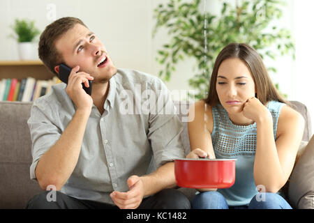 Upset couple sitting in a couch in the living room and calling insurance for home leaks Stock Photo
