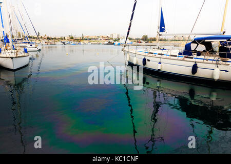 Yachts moored in Zygi harbour with fuel pollution casting a rainbow sheen on the water surface. Stock Photo