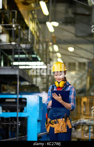 Portrait of pretty young woman wearing  hardhat looking clipboard writing something while  working in modern plant Stock Photo