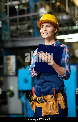Portrait of female factory worker wearing hardhat holding clipboard and smiling posing in modern workshop Stock Photo
