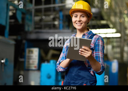 Portrait of female factory worker using digital tablet and smiling, looking at camera posing in modern workshop Stock Photo