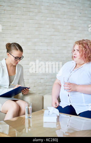 Portrait of young female psychiatrist writing on clipboard while listening to obese woman during therapy session on mental issues Stock Photo