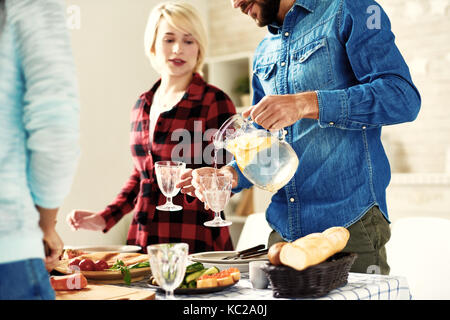 Young man pouring refreshing lemonade to glasses standing at big table with food on it while making dinner together with friends at home Stock Photo