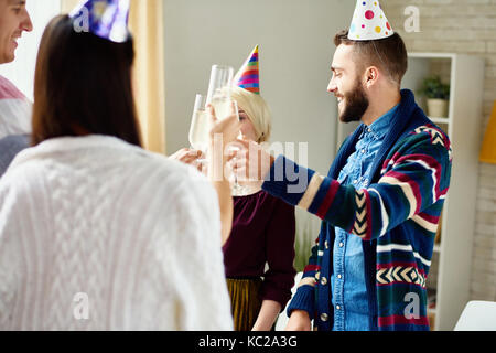 Group of happy young people wearing holiday caps celebrating Birthday with friends clinking champagne glasses during party at home Stock Photo