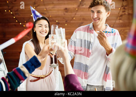 Group of happy young people wearing holiday caps celebrating Birthday with friends clinking champagne glasses during party at home, focus on smiling c Stock Photo
