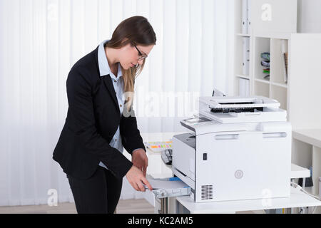 Young Businesswoman Putting Papers In Copy Machine At Workplace Stock Photo