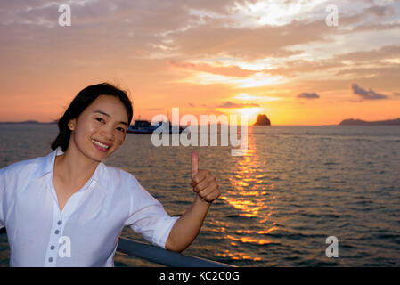 Women raise their thumb to admire the beautiful nature of colorful sky and sun at sunset over the sea on the deck of a passenger boat while cruising t Stock Photo