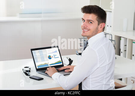 Young Happy Businessman Analyzing Financial Statistics On Laptop At Desk Stock Photo