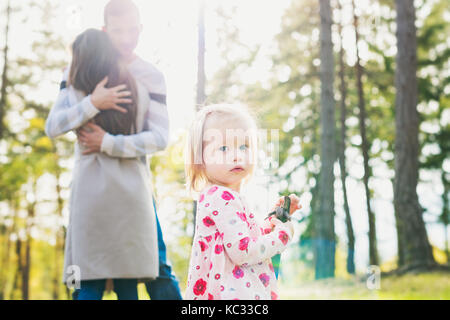 Happy young family taking a walk in a park. Parents hugging in the background with toddler girl daughter looking at camera in the foreground. Stock Photo