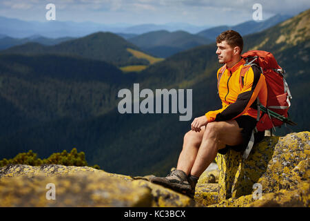 Young male hiker with backpack relaxing on top of a mountain during calm summer sunset Stock Photo