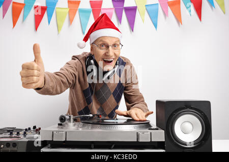 Elderly man with a christmas hat playing music on a turntable and making a thumb up sign against a wall with decoration flags Stock Photo