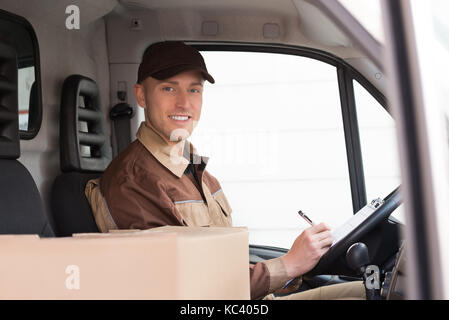 Side view of young delivery man writing on clipboard in truck Stock Photo