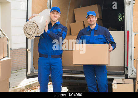 Portrait of happy delivery men carrying cardboard box and carpet outside van Stock Photo