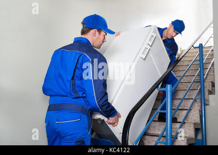 Young male movers carrying refrigerator while climbing steps at home Stock Photo