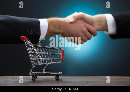 Shopping cart on table with businessmen shaking hands against blue background Stock Photo