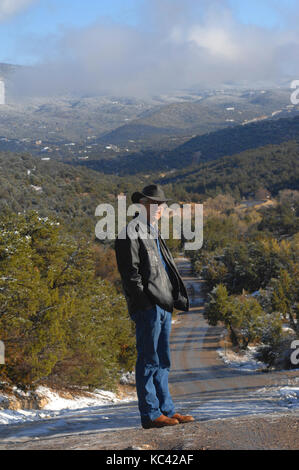 Man, dressed in blue jeans and jacket, stands at the top of a dirt road.  Road disappears into the Sandia Mountains in Tijeras, New Mexico. Stock Photo
