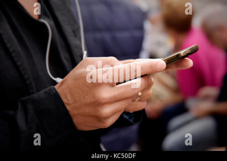closeup of a young caucasian man wearing a black jacket using his smartphone outdoors, with unrecognizable people in the background Stock Photo