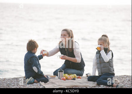 handsome mother giving a cup of hot tea to her kid with her daughter sitting beside on autumn sea backdrop Stock Photo