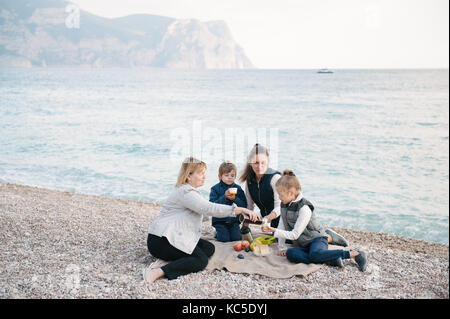 beautiful family of three generations consisting of mother, grandmother and two kids sitting on the ground near sea on weekend picnic in cold weather Stock Photo